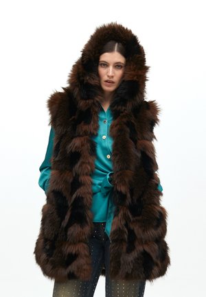Chaleco de piel sintética marrón con capucha, que presenta una mezcla de rayas marrones oscuras y claras. Llevado sobre una blusa de color teal con botones y pantalones oscuros.