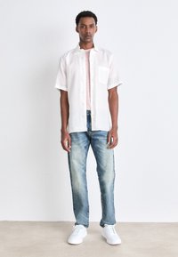Camisa de botones de manga corta blanca sobre una camiseta rosa clara, combinada con jeans azules desgastados y zapatillas blancas.