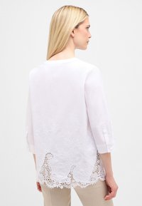 Hvid langærmet bluse med en scallop broderet kant og blomsterbroderi, lavet af letvægtsstof, med knaplukninger ved ærmerne og en simpel halsudskæring.
