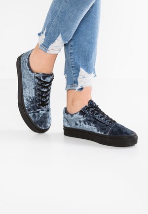 Person iført blå fløjls-sneakers med sort sål og revnede lyseblå skinny jeans mod en ensfarvet hvid baggrund.
