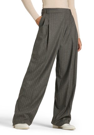 Grijze wijde pijpen broek met fijne strepen, gedragen met witte schoenen en een licht lang mouw top, één hand in de zak.