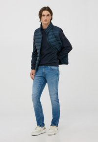 Gilet di jeans blu navy sopra un maglione blu navy, abbinato a jeans azzurri e sneakers bianche con dettagli beige. Sfondo neutro.