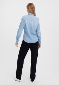 Camicia in denim azzurro chiaro con colletto a punta e polsini abbottonati, abbinata a pantaloni neri. La camicia presenta un orlo curvo e un dettano sul carré posteriore.
