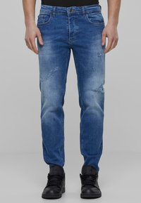 Jeans de mezclilla azul con corteSlim, presentan detalles desgastados y áreas descoloridas, complementados por un diseño clásico de cinco bolsillos. Se llevan zapatillas negras.