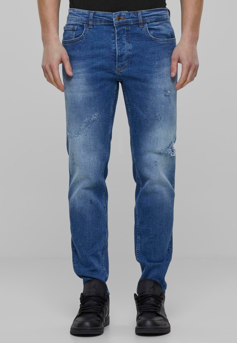 Jeans de mezclilla azul con corteSlim, presentan detalles desgastados y áreas descoloridas, complementados por un diseño clásico de cinco bolsillos. Se llevan zapatillas negras.