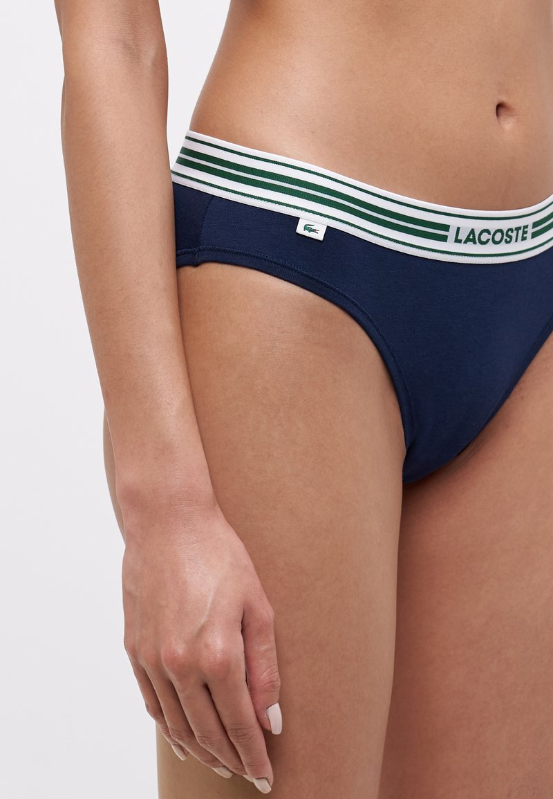 Costume da bagno blu navy con un elastico in vita a righe bianche e verdi. Presenta una piccola etichetta con il logo Lacoste sul davanti. Tessuto morbido.