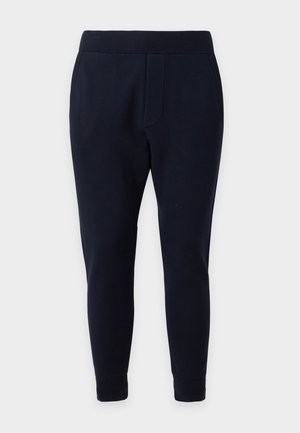Jeune homme debout portant un sweat-shirt blanc avec un texte noir, un pantalon noir et des baskets blanches avec des accents rouges et bleus.