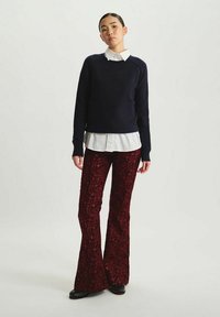 Un pull en tricot bleu marine superposé sur une chemise blanche à col, associé à un pantalon à paillettes bourgogne évasé, porté avec des chaussures noires sur un fond neutre.