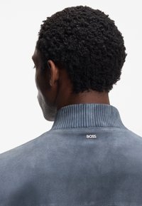 Giacca in camoscio grigia con collo a coste. Presenta un piccolo logo "BOSS" sul retro. Capelli corti e strutturati.