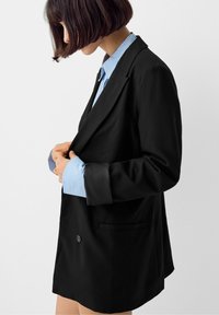 Žena upravující černý oversized blejzr s modrými a bílými pruhovanými manžetami proti bílému pozadí.