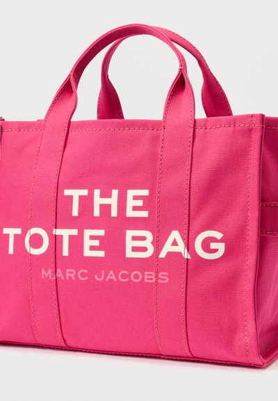 Ροζ καμβάς τσάντα tote με διπλές λαβές, διακοσμημένη με έντονο λευκό κείμενο: "Η ΤΣΑΝΤΑ TOTE" και "MARC JACOBS". Ραφές σε όλη την επιφάνεια.