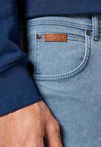 Jeans de mezclilla de color azul claro con una textura suave y costuras en contraste. Presentan un parche de marca de cuero marrón y acentos de botones metálicos.