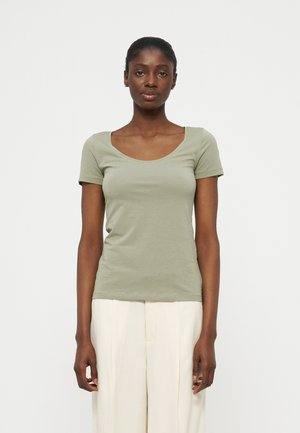 Γυναίκα που στέκεται στραμμένη προς τα εμπρός, φορώντας ένα ανοιχτοπράσινο κοντομάνικο μπλουζάκι με στρογγυλή λαιμόκοψη και λευκό παντελόνι με πλατύ πόδι, σε απλό φόντο.