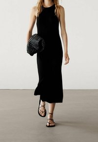 Schwarzes, geripptes Maxikleid mit hohem Ausschnitt, kombiniert mit einer geflochtenen schwarzen Handtasche und flachen Sandalen mit Riemchen. Glatte Textur und figurbetonendes Design.