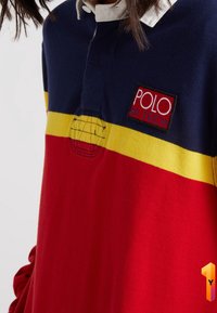 Röd och marinblå pikétröja med gula ränder, har en vit krage och en logo patch med texten "POLO HI TECH" på bröstet. Mjukt tyg.
