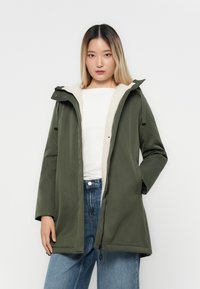 Parka vert olive avec une capuche, dotée d'une doublure en polaire crème et d'une fermeture éclair à l'avant. Le design comprend des poches latérales et des liens ajustables.