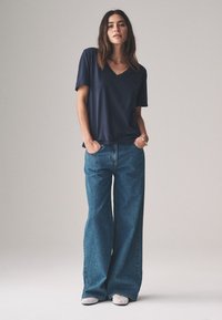 Marineblauw V-hals T-shirt gecombineerd met brede blauwe spijkerbroeken. De denim heeft een klassiek ontwerp met vijf zakken. Het model staat met handen in de zakken.