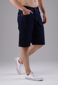 Navy blue denim shorts with five pockets, metal button, and stitching accents, worn with white low-top sneakers on a light background.