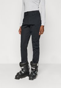 Μαύρο παντελόνι σκι από αδιάβροχο υλικό, με ζώνη με κουμπιά και εφαρμοστή γραμμή, σε συνδυασμό με μαύρα μπότες σκι.