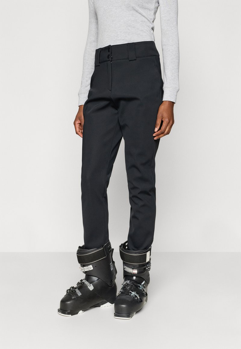 Μαύρο παντελόνι σκι από αδιάβροχο υλικό, με ζώνη με κουμπιά και εφαρμοστή γραμμή, σε συνδυασμό με μαύρα μπότες σκι.