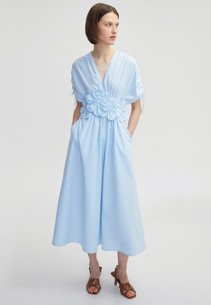Robe bleu clair avec un col en v, des manches courtes et des détails floraux à la taille. Comprend deux poches latérales et une jupe fluide.