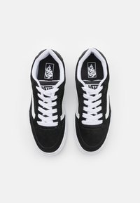 Schwarze Wildleder-Sneaker mit weißen Schnürsenkeln, ausgestattet mit einem weißen Seitenstreifen und Gummisohlen. Das Innere weist eine Markenpräsenz auf schwarzem Hintergrund auf.