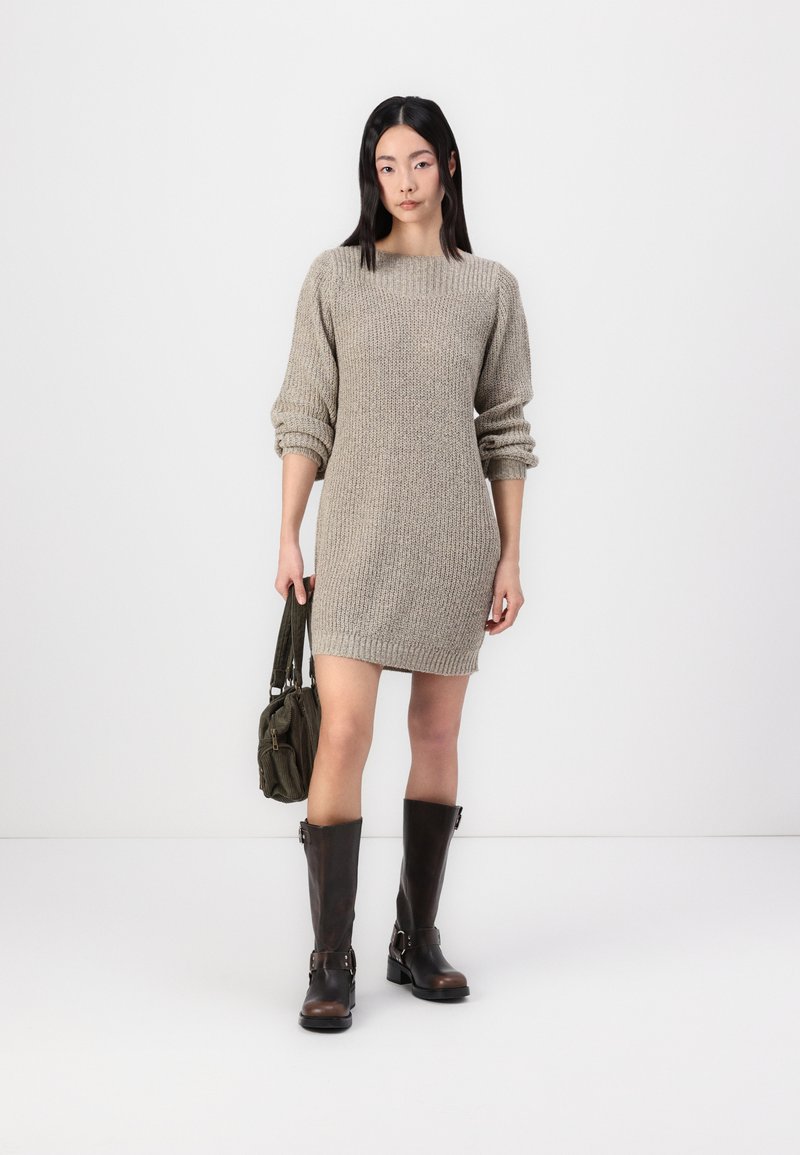 Chunky stickad, beige oversized tumlare med långa ärmar; kombinerad med mörkbruna knähöga stövlar och en grön tyghandväska.