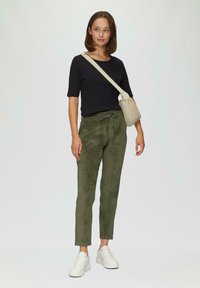 Schwarzes, geripptes Kurzarmoberteil, grüne, samtige enganliegende Hose mit Kordelzug und weiße Sneakers. Das Model trägt eine beige Tasche.