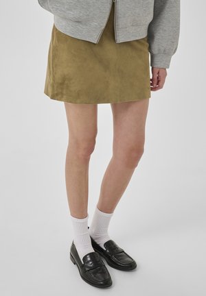 Person iført en lysebrun ruskind mininederdel, grå lynlåstrøje, hvide sokker og sorte læder loafers mod en ensfarvet baggrund.