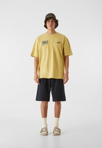 Gul bomull T-skjorte med teksten "AMALFI COAST", kombinert med marineblå shorts og brune sandaler. Modellen har på seg en grønn strikket bøttehatt og lyse sokker.