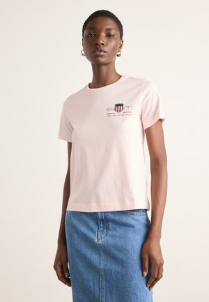 T-shirt di cotone rosa chiaro con colletto rotondo, maniche corte e stampa grafica del logo sul petto. Abbinata a una gonna di jeans blu.