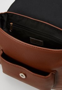 Ruskea teksturoitu nahkakäsilaukku, jossa on etutasku ja musta sisävuori, jossa on vetoketjullinen osasto ja kangasbrändin etiketti.