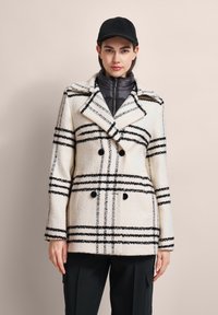 Doppelreiher-Mantel aus cremefarbener Wollmischung mit schwarzem Karomuster, mit schmalem Kragen und großen Knöpfen. Über einer grauen Zip-Jacke getragen.