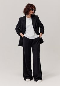 Czarna marynarka i szerokie spodnie wykonane z gładkiego materiału; zestawione z białym T-shirtem i białymi sneakersami; okulary przeciwsłoneczne dodają kontrastu.