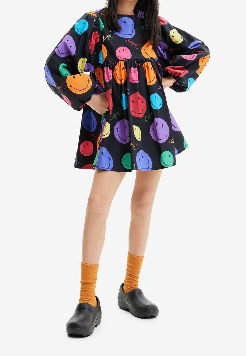 Kolorowa sukienka z dużymi wzorami uśmiechniętych buziek w odcieniach fioletu, pomarańczy, zieleni i różu na czarnym tle; zestawiona z czarnymi butami i pomarańczowymi skarpetkami.