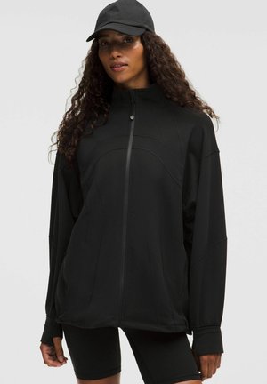 Musta tõmblukkidega jakk, mis on valmistatud pehmest kangast, kõrge kaelusega, laia lõikega ja õmbluste detailidega. Kombineeritud sobiva musta mütsiga.