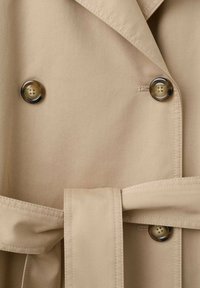 Trench-coat beige avec boutons marron foncé à quatre trous et une ceinture assortie en tissu nouée à la taille.