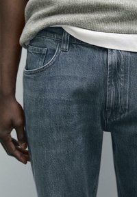 Jeans in denim di un blu medio, con un classico design a cinque tasche, leggeri segni di usura e un maglione a coste texturizzato sopra.