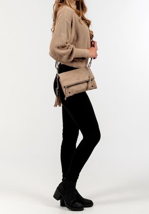 Frau trägt einen beigen Strickpullover, schwarze Hose und schwarze Stiefel mit Absatz, dazu eine beige Umhängetasche mit Quasten.