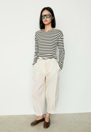 Donna che indossa una maglia a maniche lunghe a righe bianche e nere, pantaloni crema a gamba larga con cintura, ballerine marroni e occhiali da sole scuri, in piedi contro un muro bianco.