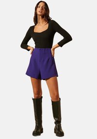 Haut noir à manches longues côtelé, décolleté carré, associé à un short violet taille haute avec des plis. La modèle porte des bottes noires jusqu'au genou.