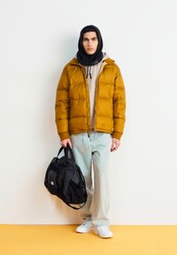 Puffer jacket in mustard, worn over a beige hoodie. Light blue jeans and white sneakers. Black duffel bag with handles.