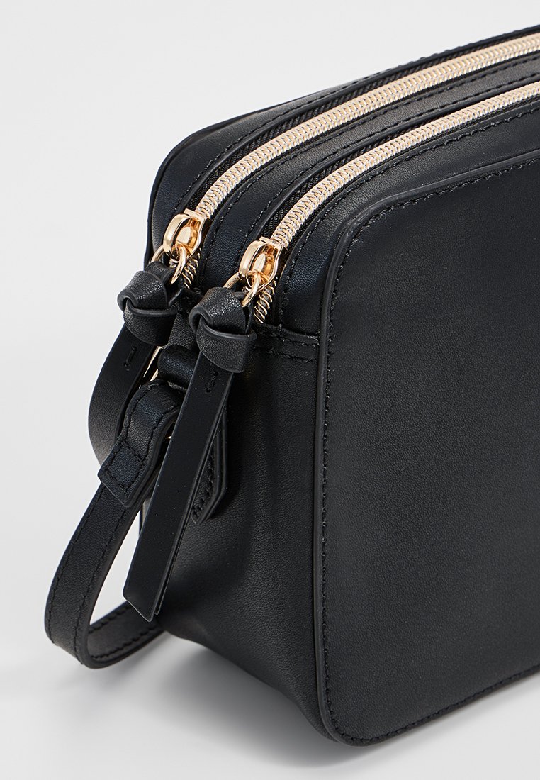 Bolso cruzado de cuero negro con dos cremalleras doradas y tiradores de cuero a juego, mostrando un detalle de la esquina y el lazo de la correa.