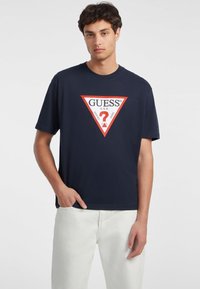 T-shirt in cotone blu navy con una grafica a triangolo rossa e bianca con la scritta "GUESS" e un punto interrogativo, maniche corte e vestibilità rilassata.