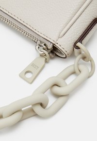 Portafoglio in pelle crema con texture e accento marrone, dotato di zip argentata e di una robusta tracolla in catena beige. Attaccato il logo DKNY.