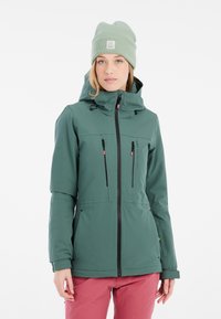 Grüne wasserdichte Jacke mit einer taillierten Passform, schwarzem Reißverschluss, zwei Vordertaschen und Kapuze; getragen mit einer hellgrünen Mütze.