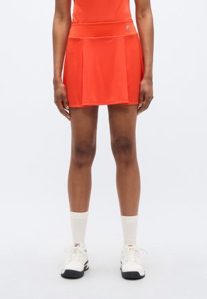 Falda de tenis plisada roja con una cintura alta y detalle de logo, hecha de tela ligera. Se combina con zapatillas blancas y calcetines de canalé texturizados.