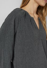 Gray textured blouse with a V-neck and gathered shoulders. Features a crinkled fabric with subtle line patterns. Loose fit and long sleeves.