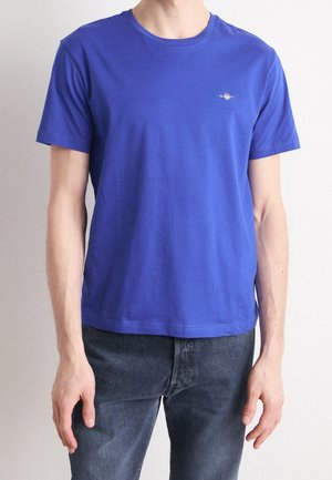 Personne portant un t-shirt bleu uni à manches courtes et col rond ainsi qu'un jean en denim foncé, debout devant un fond neutre clair.