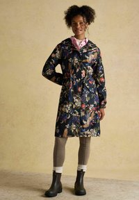 Femeie în picioare, purtând o haină închisă la culoare cu model floral, cămașă roz cu guler, colanți în carouri, șosete albe și cizme scurte închise la culoare.